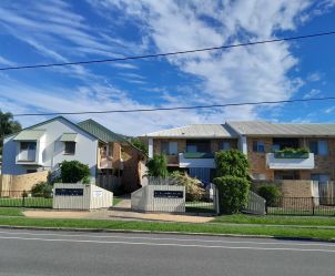 Two modern apartment buildings with brick and white facades sit under a bright blue sky with scattered clouds, surrounded by green landscaping.